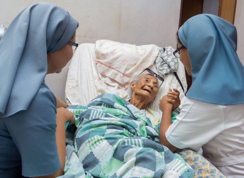 Two nuns with blue wimples comfort an elderly women in a hospital bed