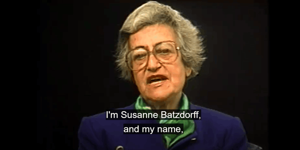 A woman with short grey hair, wearing a green scarf and navy top and glasses