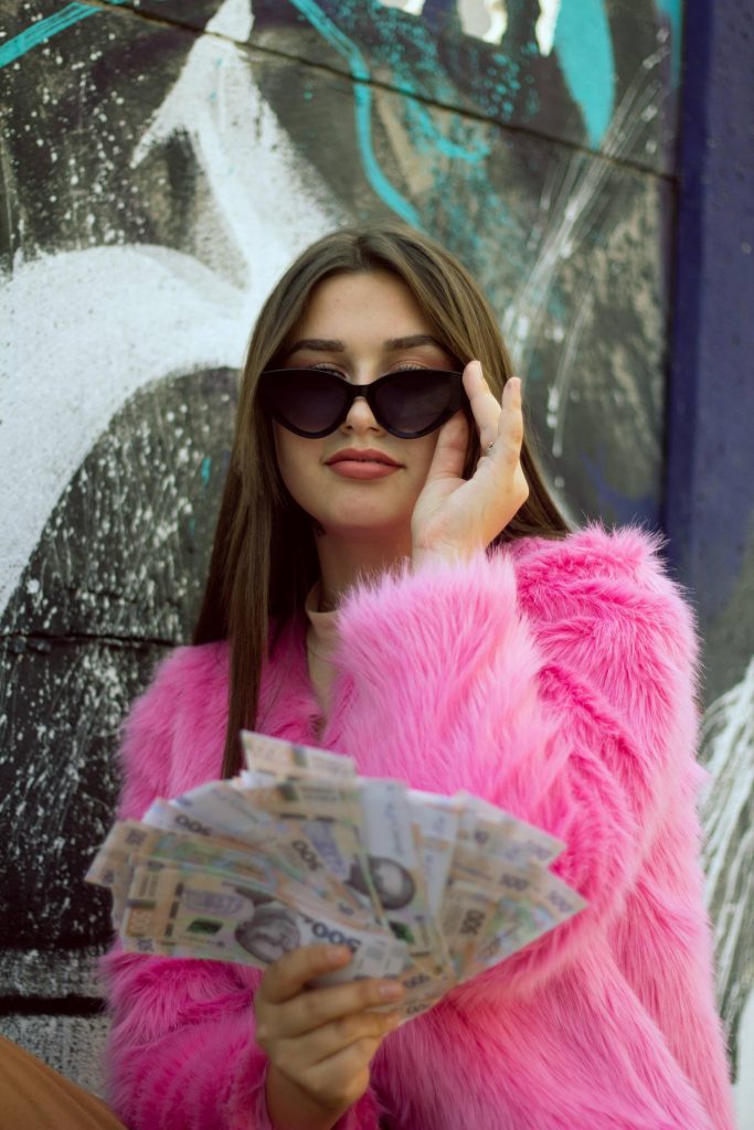 a woman in a pink feather top holds cash and looks over sunglasses
